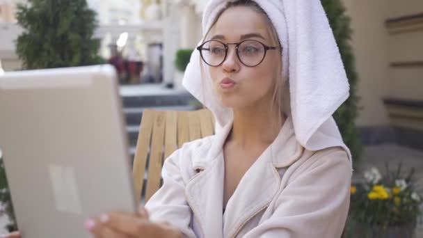 Ritratto di divertente bella giovane donna in occhiali e accappatoio con asciugamano sulla testa guardando tablet seduto sul cortile. Ragazza fiduciosa godendo giornata di sole all'aperto . — Video Stock