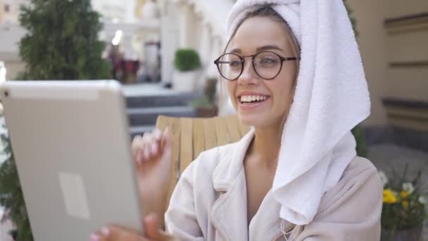 Retrato de mujer joven en gafas y albornoz con toalla en la cabeza hablando con su amiga usando la cámara en su tableta sentada en el patio trasero. Chica segura disfrutando de un día soleado al aire libre — Vídeos de Stock