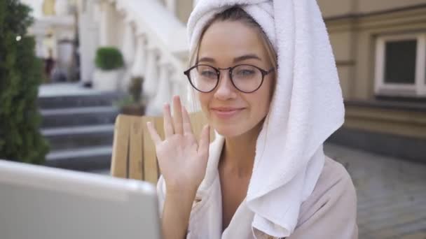 Portret cute młodej kobiety w okularach i szlafrok z ręcznikiem na głowie rozmawia z przyjacielem za pomocą aparatu na jej tablecie siedzi na podwórku. Pewna dziewczyna ciesząc słoneczny dzień na świeżym powietrzu — Wideo stockowe