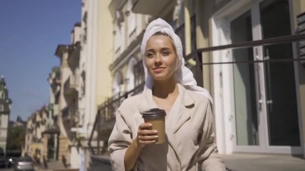 Retrato sorrindo jovem em roupão de banho com toalha na cabeça andando pela rua bebendo café. Menina confiante desfrutando de um belo dia na cidade — Vídeo de Stock