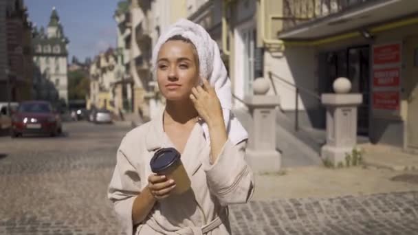 Retrato de una joven muy sonriente en albornoz con toalla en la cabeza caminando por la calle tomando café. Chica segura disfrutando de un hermoso día en la ciudad — Vídeos de Stock