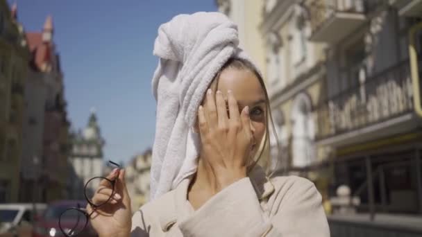 Portrait de jeune femme mignonne avec serviette sur la tête dans des lunettes regardant la caméra souriant tout en se tenant debout dans la rue. Fille confiante profiter d'une belle journée en plein air — Video