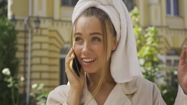 Retrato de una linda joven sonriente en albornoz con toalla en la cabeza hablando por teléfono celular en la calle. Chica segura disfrutando de un hermoso día en la ciudad — Vídeo de stock