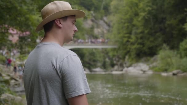 Όμορφος άντρας με καπέλο που κοιτάζει στο ποτάμι στο δάσος. Σύνδεση με τη φύση. Ταξίδια, διακοπές. — Αρχείο Βίντεο