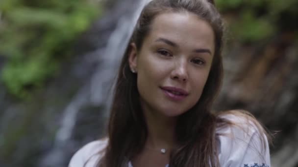 Close-up portrait of young woman with freckles on her face looking at the camera in front of waterfall. The girl spending time outdoors. Connection with nature. Slow motion — Stock videók