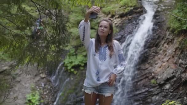Милая молодая женщина в вышитой рубашке делает селфи, стоя перед водопадом. Связь с дикой природой. Отдых на свежем воздухе, активный образ жизни. Медленное движение — стоковое видео