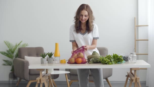 Fiatal csinos nő készül főzni egy salátát. Zöldségek és gyümölcsök az asztalon egy hangulatos konyhában. Egészséges táplálkozás, egészséges életmód. — Stock videók