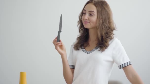 白いTシャツを着た若いイク美な笑顔の女性が、夕食を作ろうと考えてナイフを持っている。健康的なライフスタイル。食べ物を準備する気楽な主婦 — ストック動画