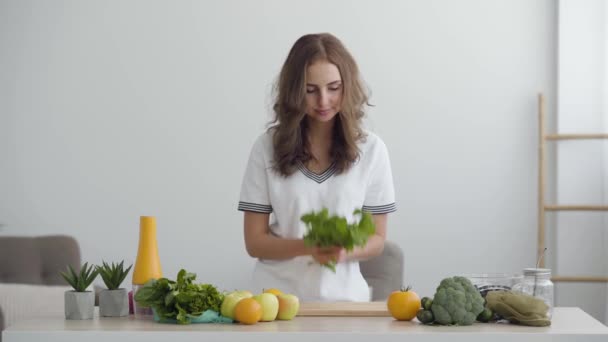Fiatal mosolygó nő szippantás friss zöldek állva az asztalnál a modern konyhában. Az egészséges táplálkozás fogalma. Nutri terapeuta, Nutraceutical, dietetikus, wellness edző. — Stock videók