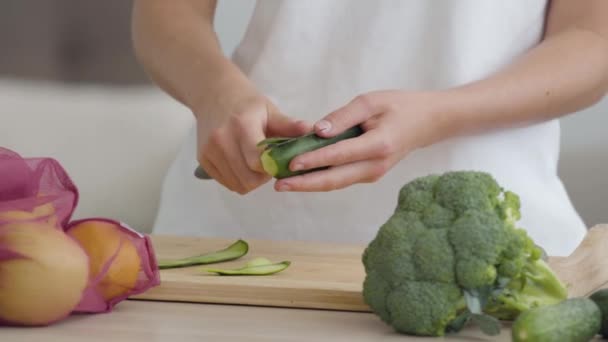 Крупный план рук юной худой женщины, чистящей огурец острым ножом за столом на кухне. Концепция здорового питания. Фрукты и овощи лежат на переднем плане — стоковое видео