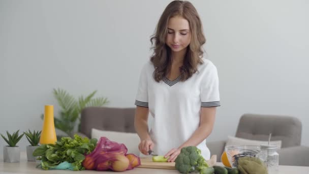 Mulher magra jovem cortando legumes com a faca afiada na mesa na cozinha. Conceito de comida saudável. Muitas frutas e legumes que se encontram em primeiro plano — Vídeo de Stock