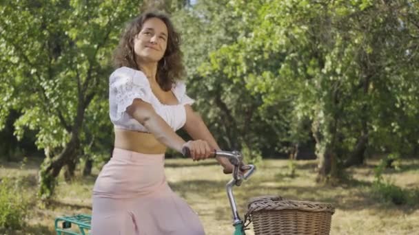 彼女の自転車で庭や公園に立って、カメラを見て巻き毛を持つかわいい若い女性。田舎の生活レトロなスタイル。美しい夏の日を楽しむ田舎の女の子 — ストック動画