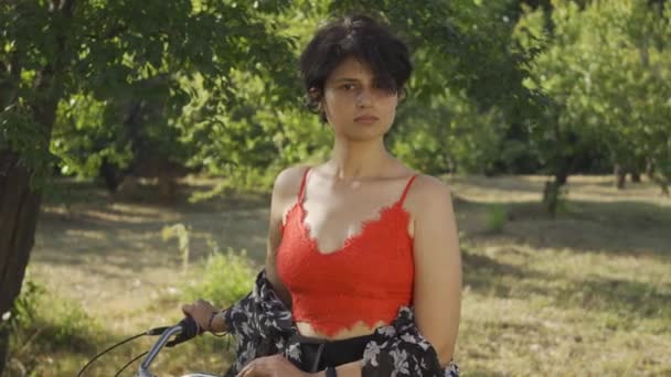 Portrait of pretty young woman with short black hair standing in the garden or park with her bicycle looking at camera. Rural life. Retro style. Country girl enjoying beautiful summer day — Stock Video