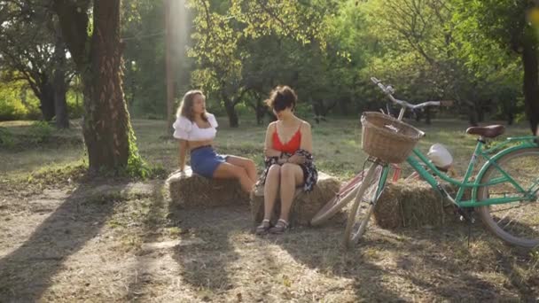 Two cute women talking while sitting on hay stacks in the summer garden, their bicycles standing near. Rural life. Retro style. Beautiful summer day — Stock Video