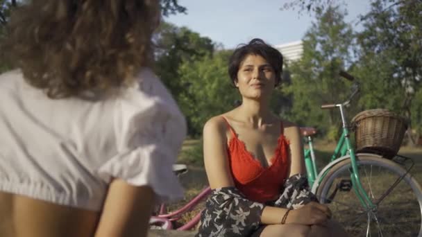 Dos mujeres lindas hablando mientras están sentadas juntas en el jardín de verano, sus bicicletas de pie cerca. La vida rural. Estilo retro. Hermoso día de verano. Disparos sobre el hombro — Vídeos de Stock