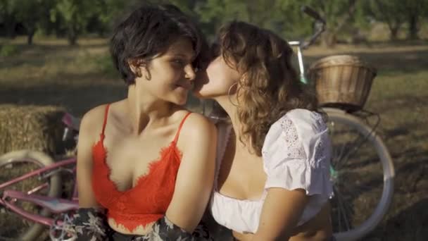 Dos mujeres lindas sentadas juntas en el jardín de verano, sus bicicletas de pie cerca. Hermosa pareja lesbiana. La vida rural. Estilo retro. Día caliente de verano — Vídeo de stock