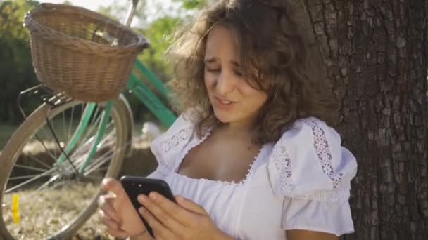 Portrait cute young woman with curly hair sitting under the tree with her phone in the garden or park, her bicycle standing near. Rural life. Retro style. Country girl enjoying beautiful summer day — Stock Video