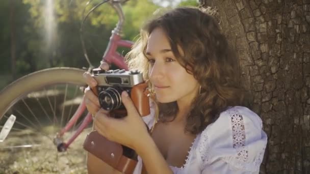 迷人的年轻摄影师卷曲的头发坐在树下拍照使用旧相机在花园或公园，她的自行车站在后面。农村生活。复古风格 — 图库视频影像