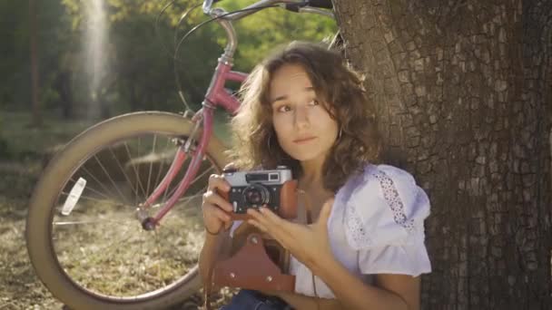 Linda joven fotógrafa con el pelo rizado sentado debajo del árbol tomando fotos usando una vieja cámara en el jardín o el parque, su bicicleta de pie detrás. La vida rural. Estilo retro — Vídeos de Stock