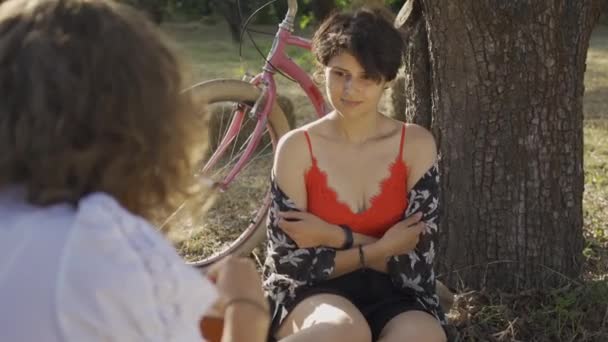 彼女の友人が庭で写真を撮っている間、短い髪を持つかわいい若い女性が木の下に座っています。後ろに立っている自転車。田舎の生活レトロなスタイル — ストック動画