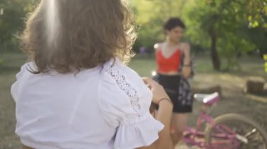 Arkadaşı bahçe veya parkta fotoğraf çekerken kısa saçlı, bisikletiyle poz veren genç beyaz bir kadın. Kırsal yaşam. Retro tarzı