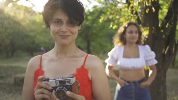 Attraktiv kaukasisk kvinna med lockigt hår poserar i bakgrunden medan hennes vän tar foto med hjälp av gamla kameran i förgrunden i trädgården eller parken. Landsbygdslivet. Retrostil. Skytte över axeln — Stockvideo