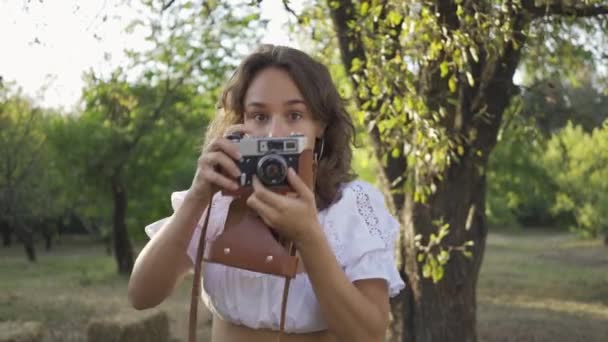 Portret cute młodego fotografa z kręcone włosy patrząc na aparat podczas robienia zdjęć za pomocą starego aparatu w ogrodzie lub parku. Życie na wsi. Styl retro — Wideo stockowe