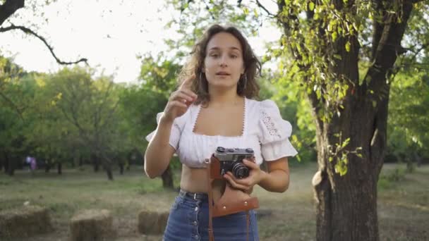 Bahçe veya parkta eski kamera kullanarak fotoğraf çekerken kameraya bakarak kıvırcık saçlı çekici genç fotoğrafçı. Kırsal yaşam. Retro tarzı — Stok video