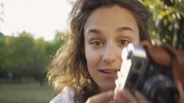 Portrait of cute young female photographer with curly hair looking at the camera while taking photo using old camera in the garden or park. Rural life. Retro style — Stock Video