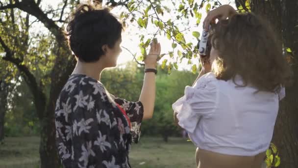 Entzückende junge Fotografin mit lockigem Haar, die das Mädchen im Garten oder Park fotografiert. die Fotografin, die ihrem Kunden hilft, besser zu posieren. Retro-Stil. Beruf — Stockvideo