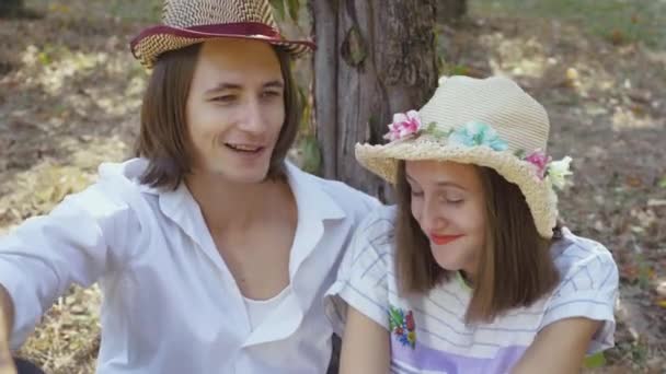 Cute couple sitting under the tree in the park talking and smiling. The man with long hair and pretty woman spending time together outdoors. Retro style. — Stock Video