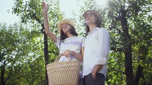 Cute couple standing under the trees in the park or garden talking and laughing, girl showing up. The man and woman in hats spending time together outdoors. Retro style. — Stock Video