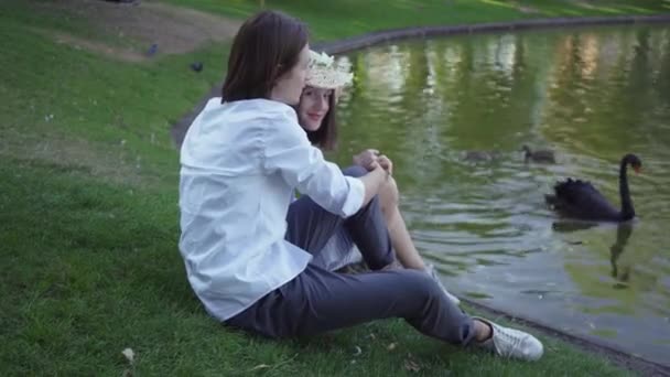 Счастливая пара сидит на берегу озера или реки, разговаривает и улыбается. Молодой кавказский мужчина и женщина проводят время вместе в парке или саду. Вид сбоку . — стоковое видео