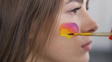 Genç ve güzel bir kadının yakın çekim portresi. Sanatçı boyama sosyal medyanın çok renkli simgesi kızlar üzerinde küçük yumuşak fırça kullanarak yüz. Vücut sanatı konsepti. İnsan vücudunda sanat eserleri. Yan görünüm.
