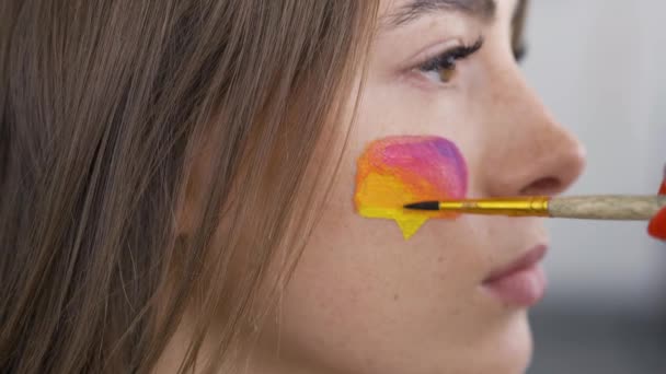 Närbild porträtt av en ung vacker kvinna. Konstnär målning Multicolor ikon av sociala medier på flickorna ansikte med hjälp av små mjuka borste. Body Art koncept. Konstverk på en människa förkroppsligar. Side View. — Stockvideo