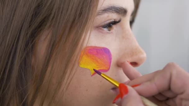 Retrato de cerca de una joven agradable. Artista está pintando cuadro de mensaje multicolor en la cara de las niñas con un cepillo pequeño. Concepto de redes sociales. Vista lateral . — Vídeos de Stock