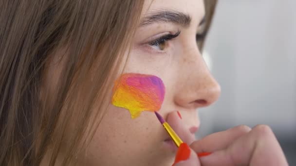 Close-up portrait of young pleasant woman. Artist is painting multicolor message box on the girls face using small brush. Social media concept — Stock Video