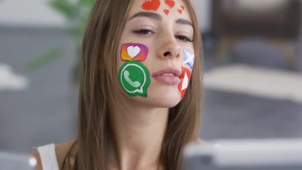 Κοντινό-up πορτρέτο της νεαρής χαριτωμένο γυναίκα με ζωγραφισμένα εικόνες μέσα κοινωνικής δικτύωσης στο πρόσωπό της κρατώντας το τηλέφωνο και το δισκίο στα χέρια. Όμορφο κορίτσι που παίρνει selfie. Εθισμός σε gadget. — Αρχείο Βίντεο