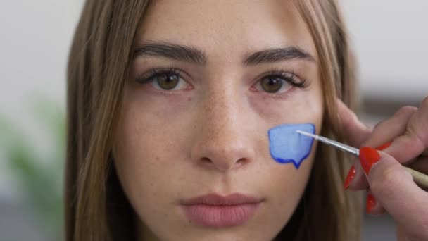 Primer plano retrato de la joven mujer agradable mirando a la cámara mientras artista pintura cero en el cuadro de mensaje en la cara de las niñas con un cepillo pequeño. Concepto de redes sociales . — Vídeo de stock