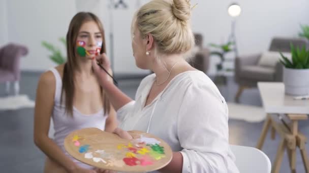 Pintura artista femenina en la cara de las niñas utilizando un pequeño cepillo suave, luego girando y mirando a la cámara. Concepto de arte corporal. Modelo posando para artista. Proceso de trabajo — Vídeos de Stock