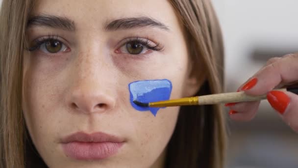 Gros plan portrait de jeune femme agréable regardant la caméra tandis que l'artiste peint avec blanc sur boîte de message bleue sur le visage des filles à l'aide d'un petit pinceau. Concept de médias sociaux . — Video