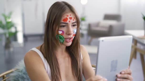 Portrait de jeune femme séduisante avec des icônes peintes sur les médias sociaux sur son visage parlant avec son ami en utilisant un appareil photo sur sa tablette. Addiction aux gadgets — Video