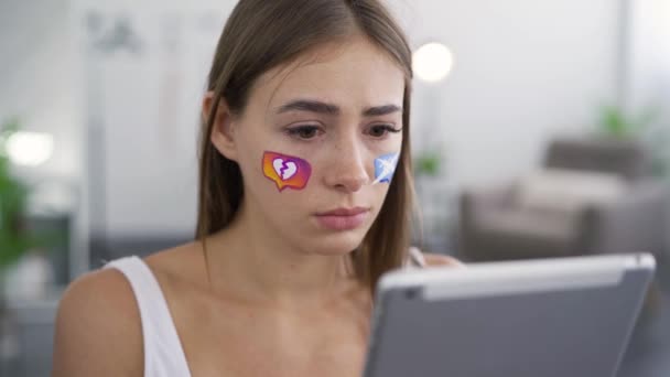 Retrato de una mujer joven sosteniendo la tableta en las manos y mirando a la cámara con tristeza. Iconos de redes sociales pintados en la cara de las niñas. Concepto de redes sociales. Adicción a los artilugios. Emociones, depresión . — Vídeos de Stock