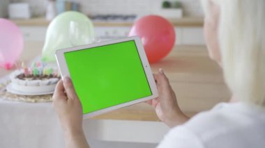 Yeşil ekranlı beyaz tablet tutan olgun kadının elleri. Doğum günü kutlaması. Kromatkey. Omzundan ateş etme