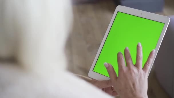 Yeşil ekranlı beyaz tablet tutan bir kadının elleri. Kadın oturuyor ve tableti izliyor. Sarışın kadın alet kullanıyor. Omzundan ateş etme. — Stok video