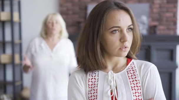 Retrato de mulher jovem em primeiro plano, sua mãe gesticulando emocionalmente no fundo. Uma jovem a discutir com a mãe. Relacionamento familiar difícil . — Vídeo de Stock