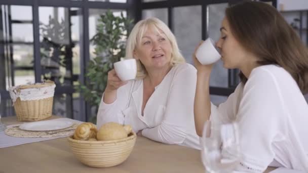 Aantrekkelijke rijpe vrouw zittend in de keuken met haar dochter drinken thee of koffie. Het meisje deelt haar geheimen met mam. Gelukkige vriendelijke familie — Stockvideo