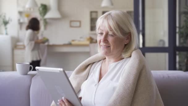 Portrait de femme mûre couverte d'une couverture assise sur le canapé regardant des photos sur la tablette tandis que sa fille nettoie la salle. Famille amicale, loisirs à l'intérieur — Video