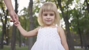 Beyaz elbiseli küçük bir kız parkta ya da ormanda yetişkin kadının elini tutarak ve elini sallayarak ayakta. Mutlu bir aile. Annelik, ebeveynlik, çocukluk.