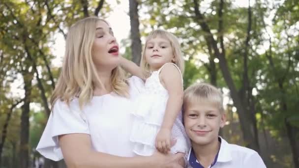 Jolie femme blonde tenant bébé fille dans ses bras tandis que le fils aîné étreignant la mère souriante. Portrait de famille heureuse avec deux enfants regardant la caméra — Video